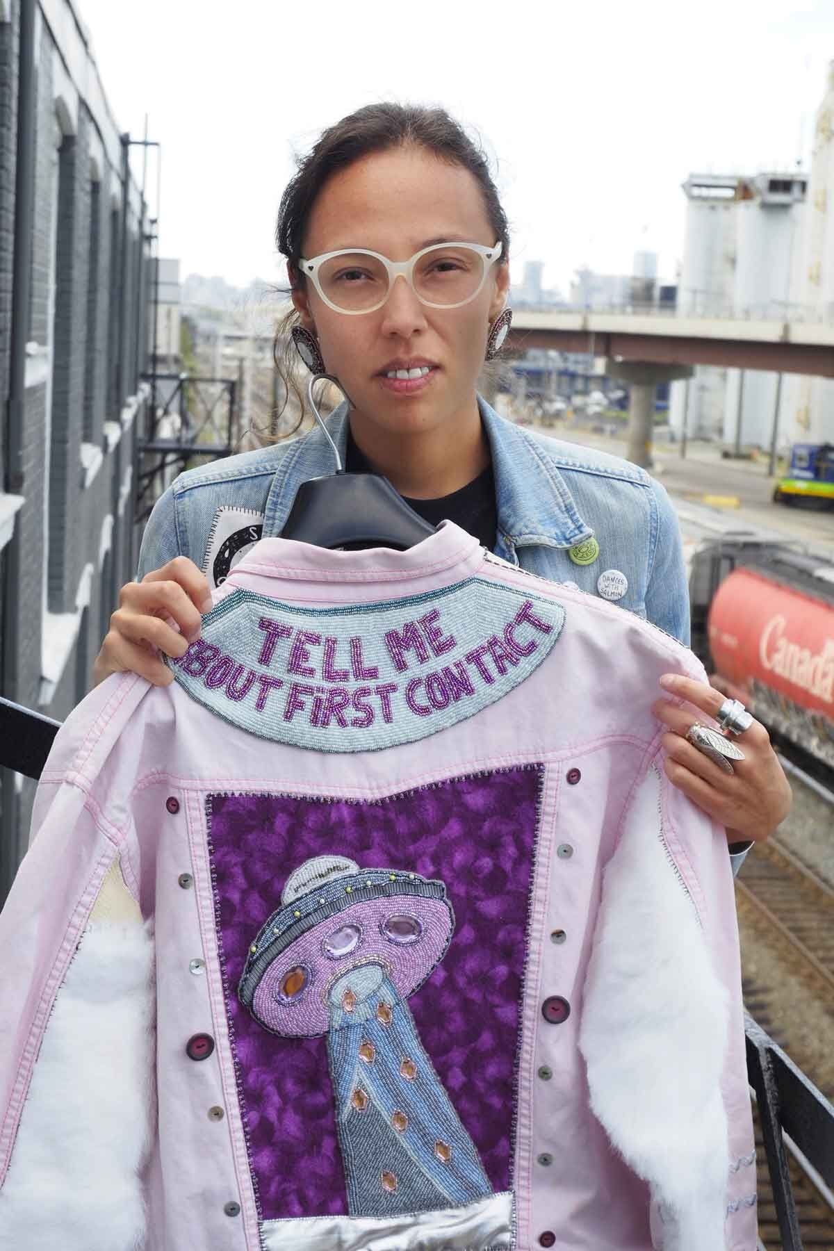 Whess Harman weating glasses stares directly into the camera holding a pink leather jacket that has "TELL ME ABOUT FIRST CONTACT"embroidered in pink and blue sequins on the upper shoulder panel of the jacket and a UFO with an abduction beam similarly embroidered just beneath