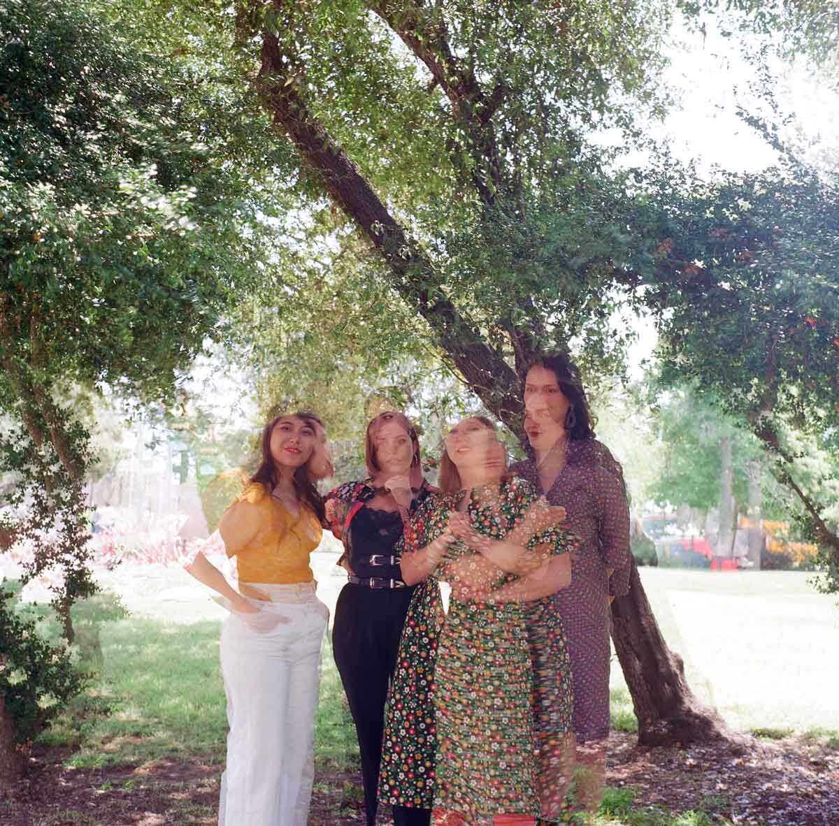 four women standing in the middle bottom of the image in front of a tree leaning diagonally from the bottom right corner to the top left. the image is slightly out of focus giving the impression of multiple images gauzily overlaid on top of each other.