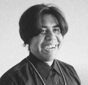 b&w photograph; Patrick Kelly, AKA Hope, looks into the bottom right corner of the frame laughing. his mid length hair is parted down the middle and flops over this round cheeks. he is wearing a dark shirt buttoned all the way up with a long beaded necklace falling out of frame over this chest.