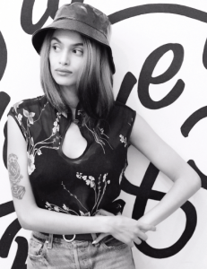 Betty Mulat, AKA Venetta, leans against a white wall with geometric squiggles, looking out the left side of the photo. she's wearing a shiny leather bucket hat over straight shoulder length hair. her left arm is perched on her hip and her right arm reaches across her waist so clasp her wrist. she is wearing a sheer black shirt with a mandarin collar over a black bra and high waisted light wash jeans accented by a black belt with a round silver buckle.