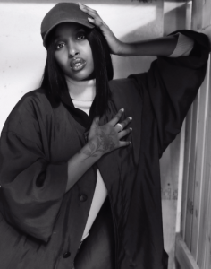 Samira Warsame, AKA ZamZam, leans against a closed panel door. her waist is in the bottom left corner of the image and her body stretches to the top right with her elbow leaning agains a white panel door, her hand tilting her baseball cap clad head to the upper left corner. she is wearing a chunky white turtleneck under a loose satin jacket. her right hand is placed over her heart.