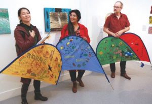 three figures stand in front of a gallery wall holding segments of tents that are painted with small vignettes of landscapes and greetings in many languages. the two right-most figures are wearing glasses and have their heads turned towards the left-most figure who has their right hand on their chest and appears to be addressing an audience behind the photographer.