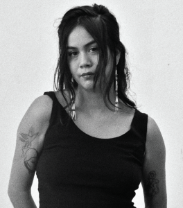 b&w photograph. Paisley Nahanee is centered and wearing a black tank top revealing a delicate tattoos on both her upper arms. her right shoulder is slightly raiser and her hair and a pair of long beaded earrings framer her face and hit her collar bones. her hair is parted down the middle and is pulled back from her face in a half-up half-down style with waved bangs covering her cheeks. her head is tilted slightly down and to the left. she makes direct eye contact with the viewer. very mona lisa esque.