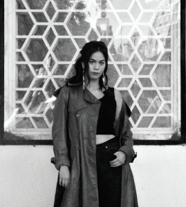 Paisley Nahanee stands in front of a window with a strong geometric design. she is wearing a black tank top, black jeans, and a brown leather trench coat. the coat is shrugged off her left shoulder and her left hand rests on her jean pocket. her head is tilted slightly down and to the right. her dark hair is in a half-up-half-down style with wavy bangs and long beaded earrings framing her cheeks.