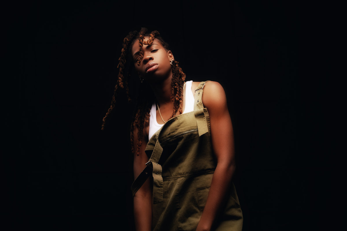 portrait photo of Haviah Mighty standing in front of a black backdrop in a white tank top and olive green overalls by Yung Yemi