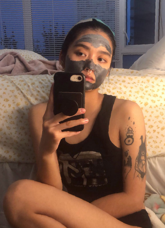 selfie courtesy of Beni Xiao of them with a face mask on, sitting in front of their bed that has a yellow floral patterned bedsheet on it.